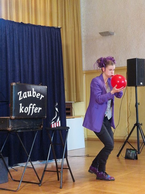 Zauberin mit lila Jacke auf der Bühne mit dem Zauberkoffer und einem roten Luftballon , der gerade aufgeblasen wird