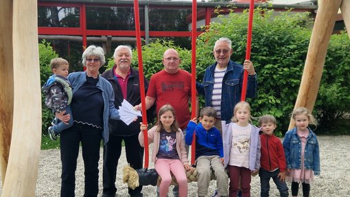 Die im Text erwähnten Personen stehen um eine massiver Holzschaukel herum. Geldumschläge werden überreicht. Kinder sitzen auf Schaukel.