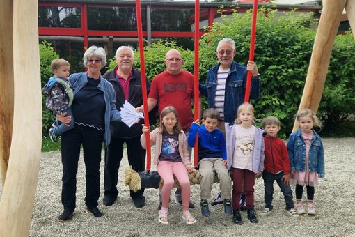 Die im Text erwähnten Personen stehen um eine massiver Holzschaukel herum. Geldumschläge werden überreicht. Kinder sitzen auf Schaukel.