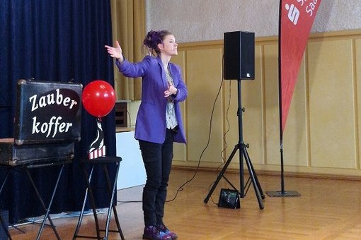 Zauberin mit lila Jacke auf der Bühne mit dem Zauberkoffer und einem roten Luftballon